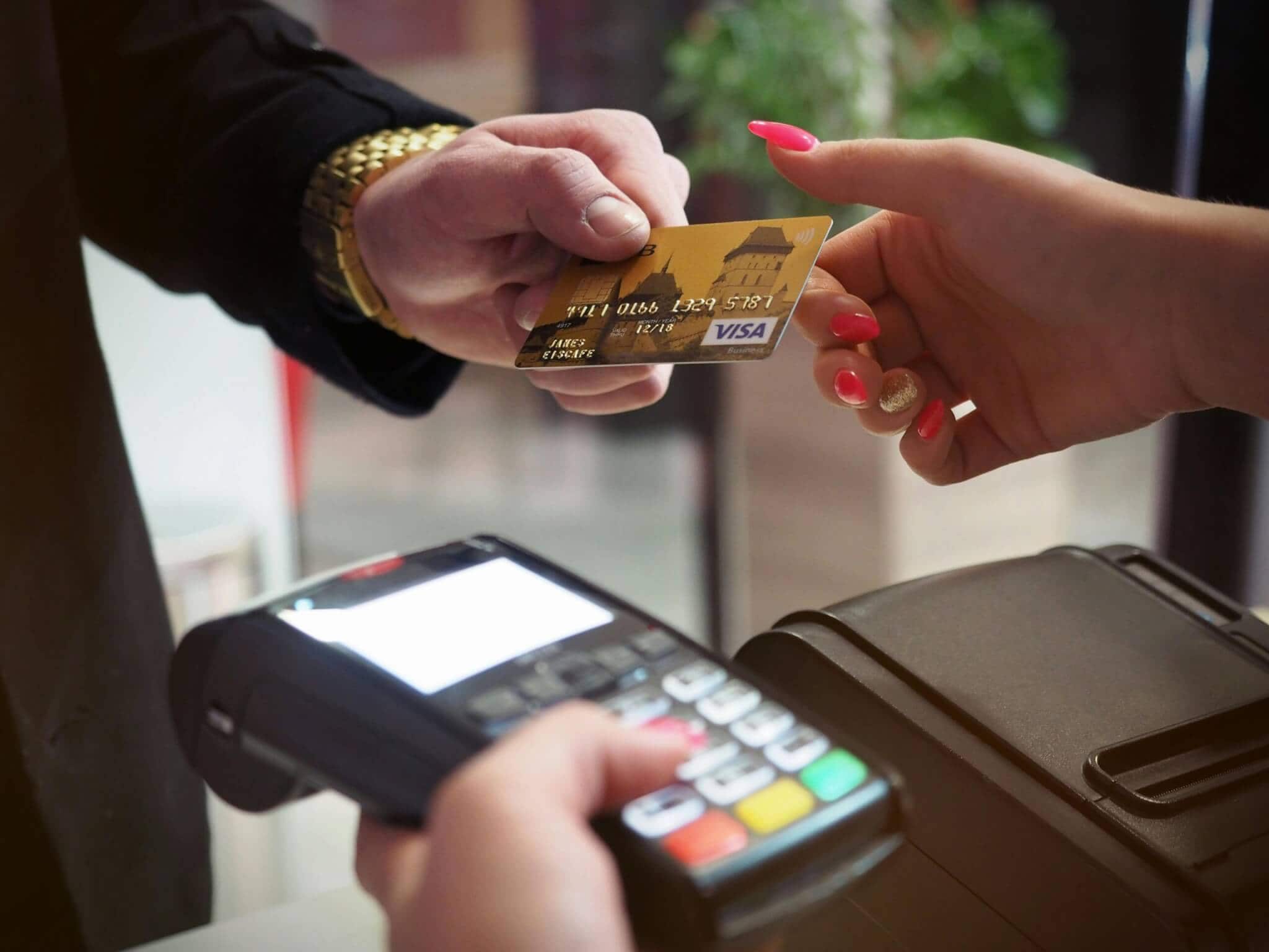 A person handing over a Visa credit card to another individual holding a card reader, ready to process a payment at a point-of-sale terminal.