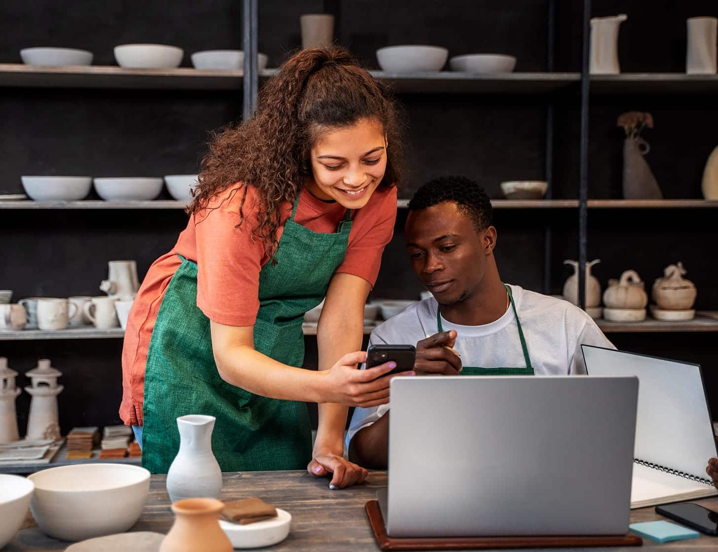 Two pottery studio owners collaborating at a worktable with ceramics, a laptop, and a smartphone
