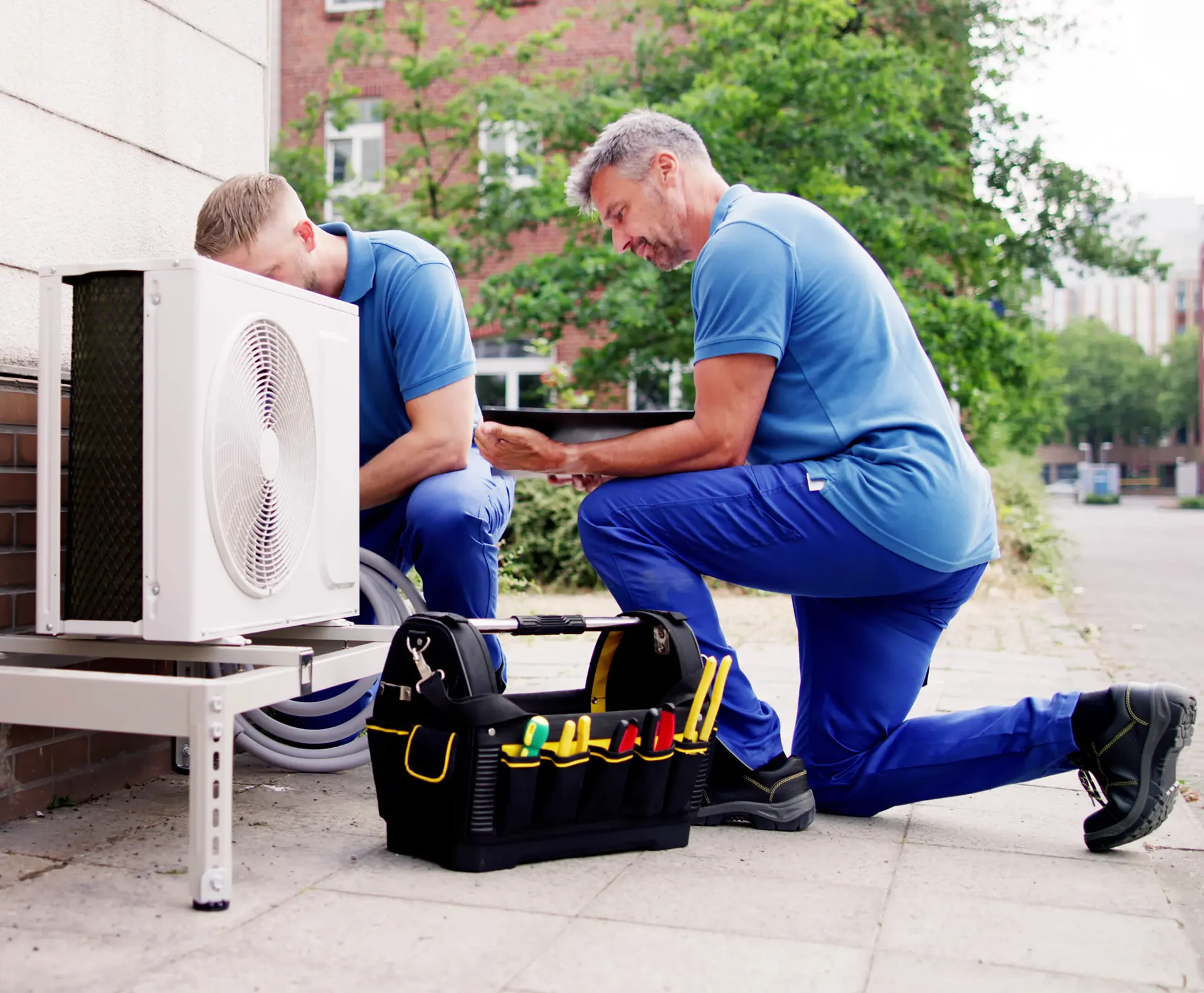 Two HVAC technicians in blue uniforms servicing an outdoor air conditioning unit