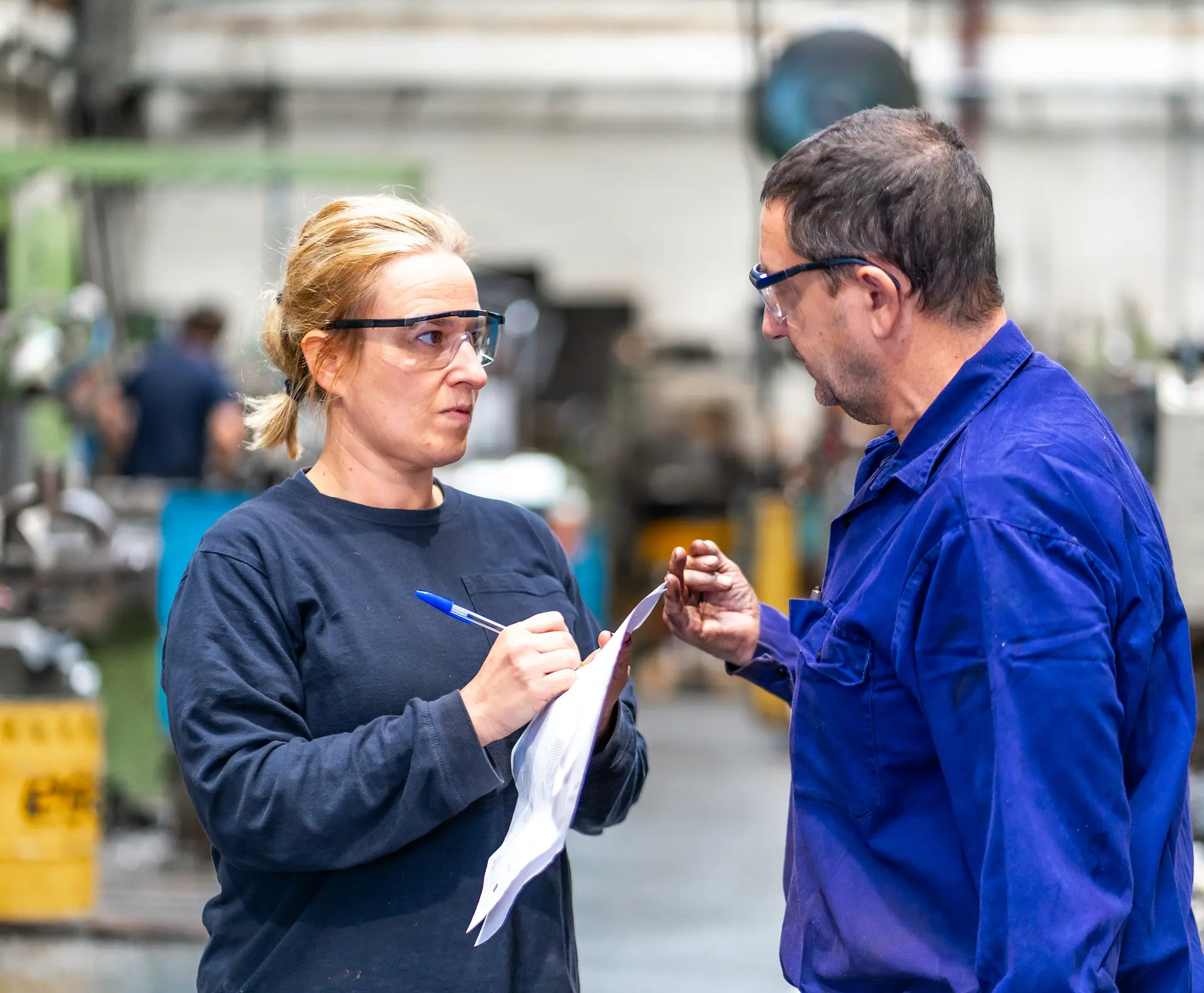 Two factory workers in safety glasses reviewing paperwork on a manufacturing floor