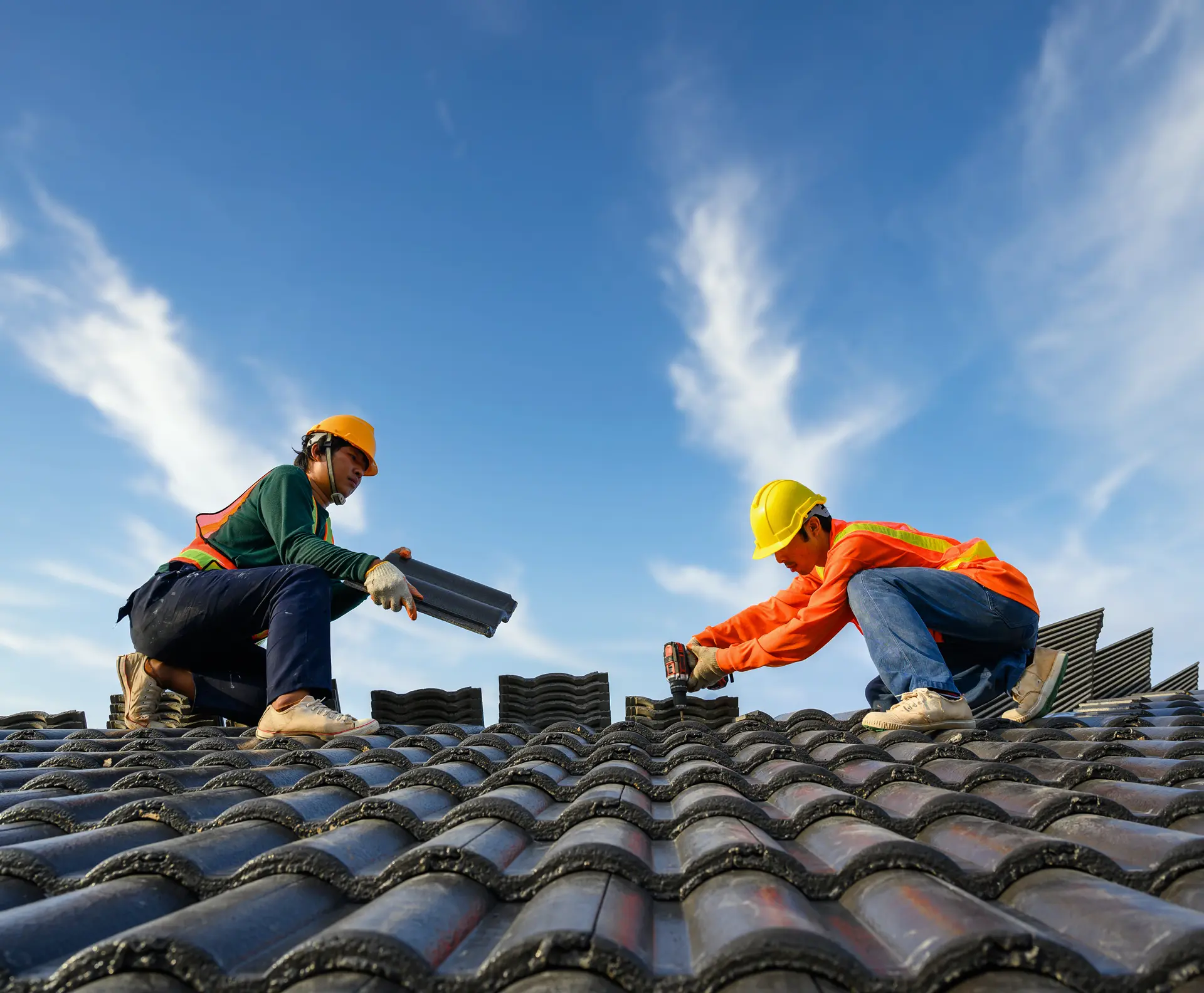 Two roofers in hard hats and safety vests installing dark roof tiles under a clear blue sky
