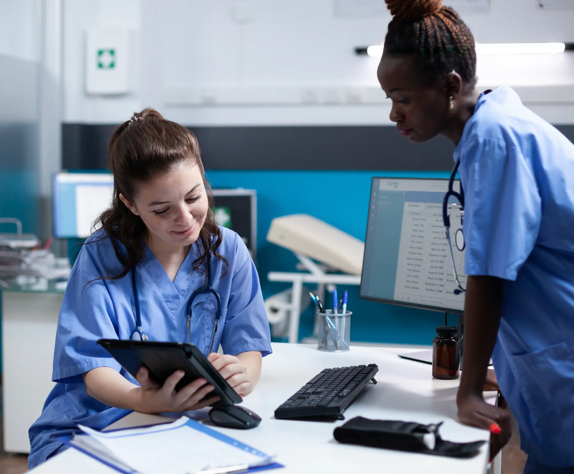 Two medical professionals in blue scrubs reviewing a tablet together in a clinic office