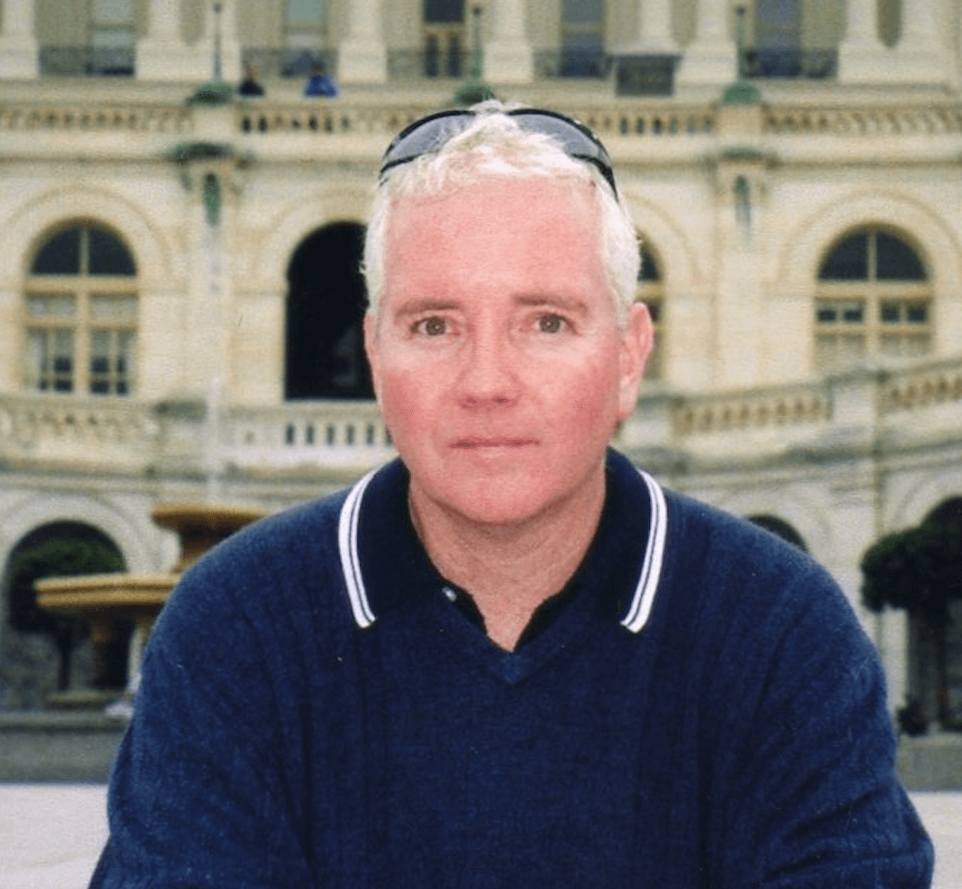 Man with short blond hair and sunglasses on his head wearing a navy sweater, standing outdoors in front of a classical building