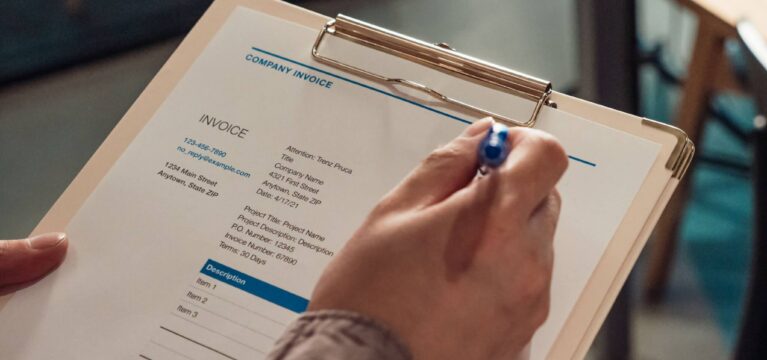 Person's hand holding a company invoice on a clipboard with a pen.