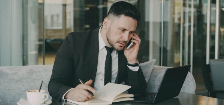 Businessman talks on phone while writing notes in a modern cafe.