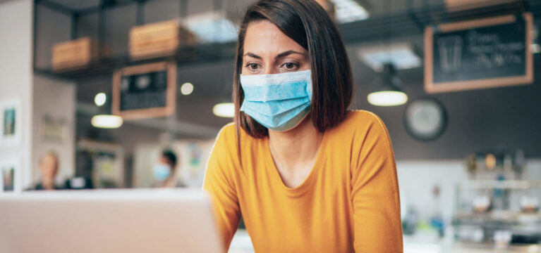 Young woman using laptop at cafe wearing face protective mask