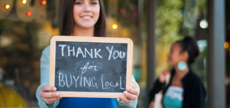 Young business owner holding a "Thank You for Buying Local" sign in front of her store with shopper in the background