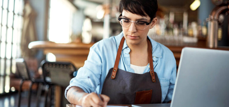 Shot of a young woman using a laptop and making notes on a clipboard at a bar