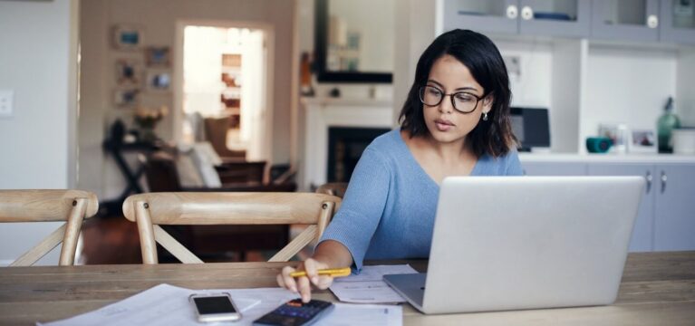 Woman sitting at home at laptop calculating expenses on the calculator in her smartphone