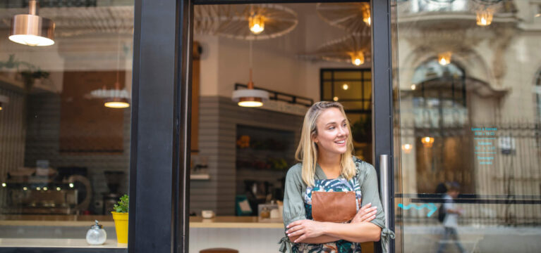 Hopeful Argentine female cafe owner standing in apron with arms crossed at open front door and looking away from camera