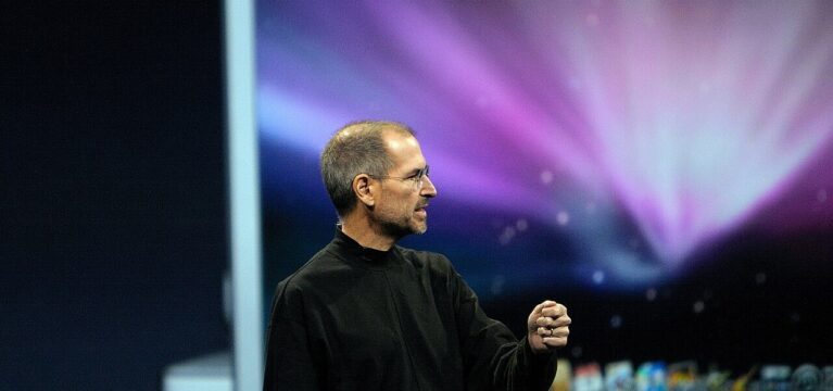 Steve Jobs delivering a presentation in his signature black turtleneck, standing against a colorful, blurred background, gesturing as he speaks.