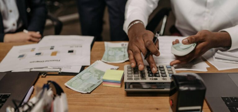 Businessperson calculating finances at a desk, surrounded by cash, documents, a calculator, and office supplies, representing financial planning and business financing decisions.