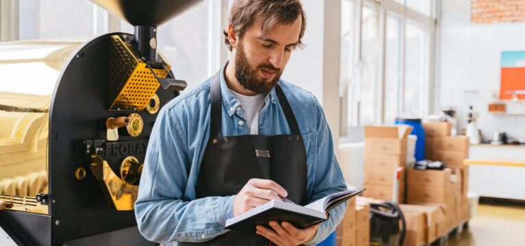 A small business owner wearing an apron stands in front of a commercial coffee roaster, writing notes in a notebook. The background includes industrial equipment and packaging materials, suggesting a coffee roasting facility.