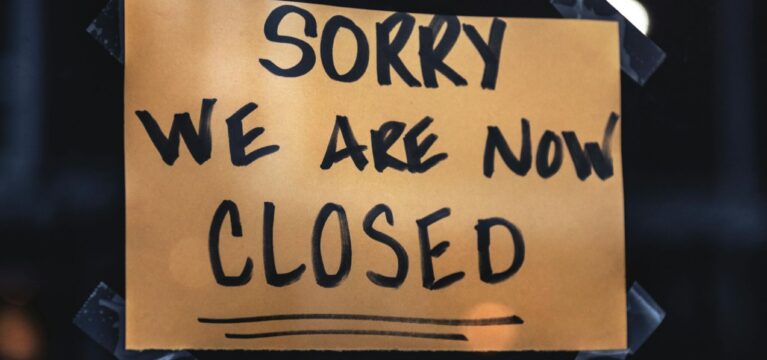 Handwritten sign on orange paper that reads "Sorry we are now closed," taped to a dark window or surface.