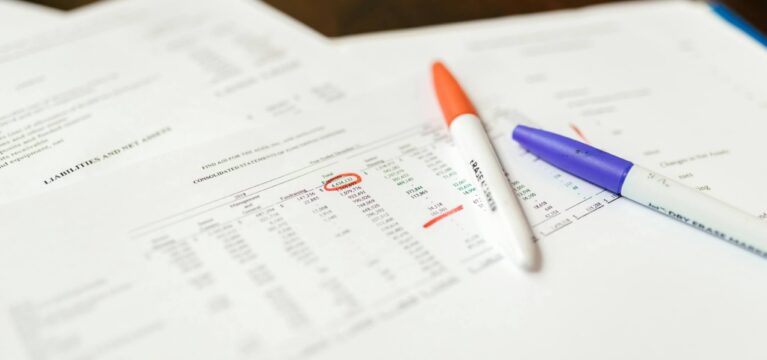 Financial documents with highlighted figures, accompanied by an orange and a blue marker, placed on a wooden desk.