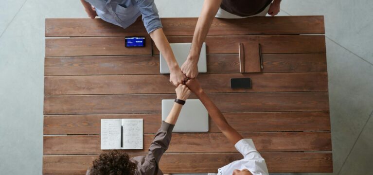 A top-down view of four business professionals fist-bumping over a wooden table, symbolizing teamwork and collaboration. The table is equipped with laptops, a notebook, and a smartphone.
