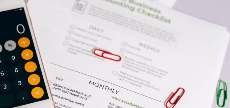 Close-up of a small business accounting checklist with a smartphone calculator, emphasizing daily, weekly, and monthly financial tasks.
