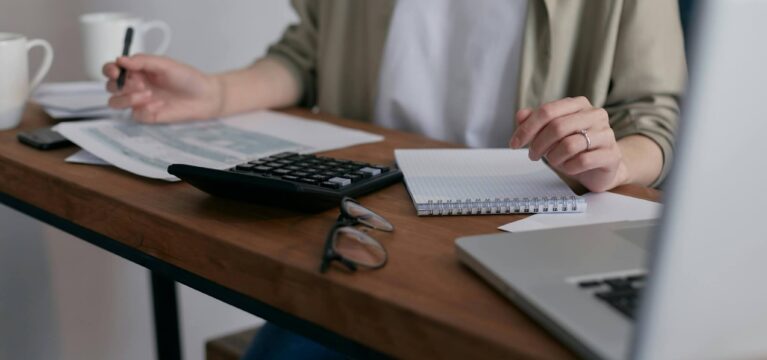 Person reviewing financial documents with a calculator, notebook, and laptop at a wooden desk.