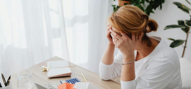 A stressed woman sitting at a desk with her head in her hands, surrounded by receipts, bills, and cash, with a laptop, calculator, and notebook on the table, symbolizing financial difficulties.