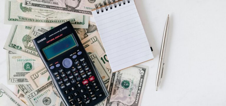 A calculator, a blank notepad, and a pen placed on a table surrounded by various U.S. dollar bills, symbolizing financial calculations and planning.