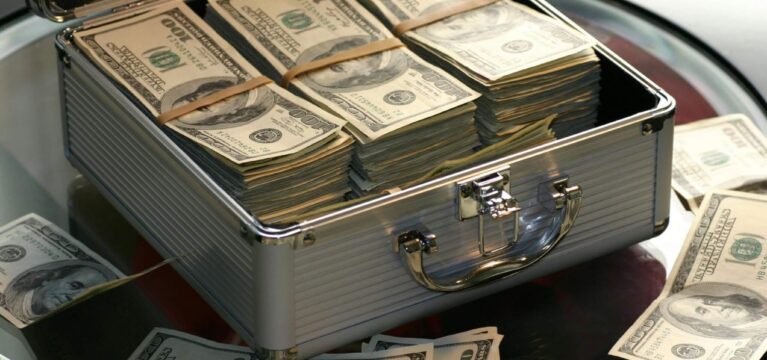 A silver briefcase filled with neatly stacked bundles of U.S. hundred-dollar bills, with additional loose bills scattered around on a glass surface.
