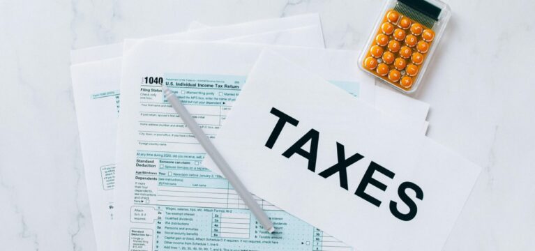 U.S. tax documents, including a 1040 individual income tax return form, placed on a white surface with a pen, a small orange calculator, and a paper labeled 'TAXES.