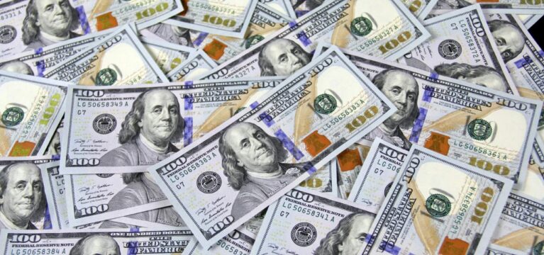 A scattered pile of U.S. $100 bills featuring Benjamin Franklin, showcasing currency details such as security strips and watermark features.