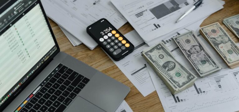 A workspace with a laptop displaying financial data, a smartphone calculator showing "3,254," printed financial reports, a pen, and stacks of US dollar bills on a wooden desk.