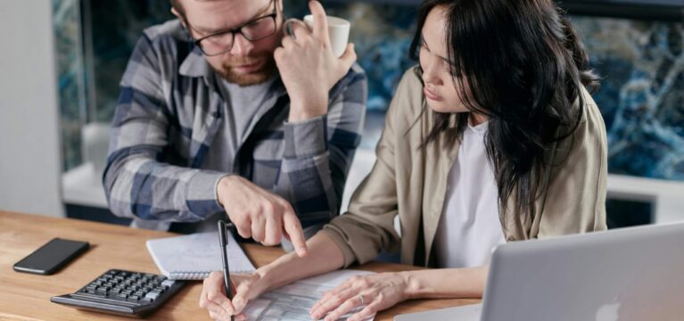 A man and a woman sitting at a wooden table, reviewing financial documents together. The man, wearing glasses and a plaid shirt, is pointing at the paperwork while the woman, dressed in a beige blazer, takes notes with a pen. A calculator, notebook, smartphone, and laptop are also on the table, indicating financial planning or budgeting.