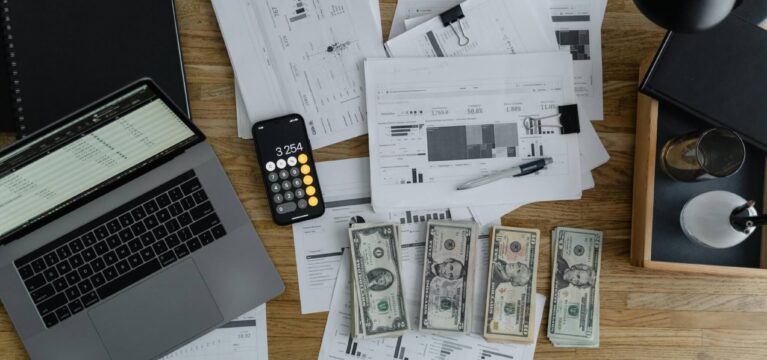 A wooden desk covered with financial documents, charts, and reports, along with a laptop, a smartphone displaying a calculator, a pen, and stacks of cash. The setup suggests financial analysis, budgeting, or cash flow management.