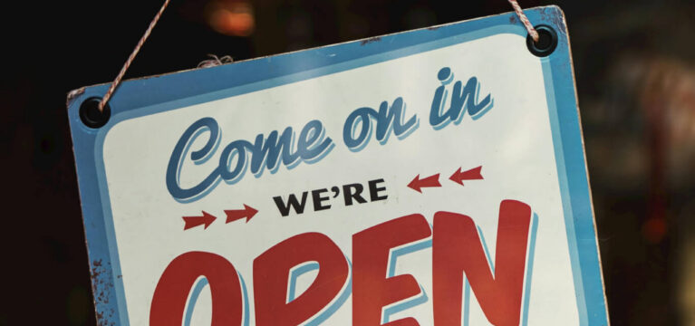 A vintage-style "Come on in, we're OPEN" sign hanging on a glass door, with "PULL HANDLE" written below. Warm lighting and reflections of indoor lamps can be seen in the background, indicating a welcoming business or storefront setting.
