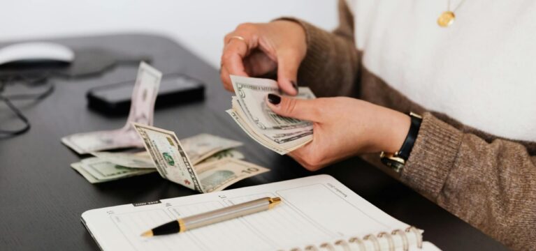 A person sitting at a desk counting U.S. dollar bills with a planner and pen in the foreground. A smartphone and computer mouse are also on the desk. The individual is wearing a white and brown sweater with a gold necklace.