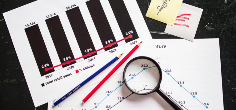 A financial analysis setup featuring printed sales charts with bar and line graphs, a magnifying glass, and red and blue pencils on a dark surface. Sticky notes with dollar symbols and upward trend drawings are also present.
