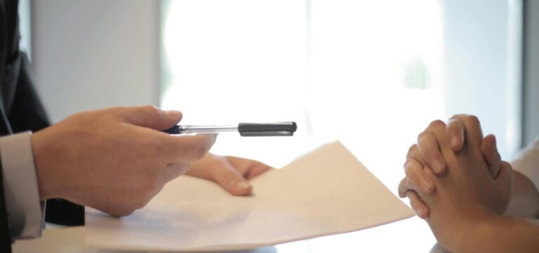A person in a business suit handing a pen and loan documents to another individual with clasped hands, symbolizing a loan agreement or financial transaction.