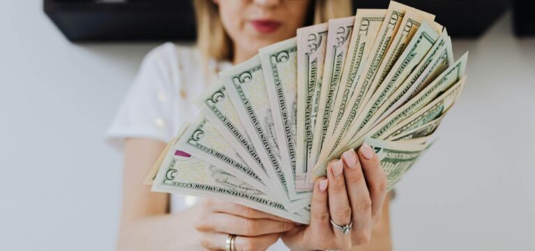 A woman wearing glasses holds a large fan of U.S. dollar bills in her hands, displaying various denominations.