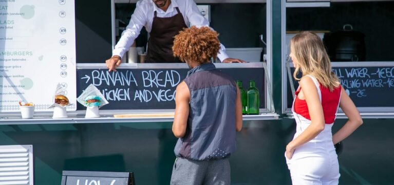 A food truck vendor in a white shirt and brown apron points to a chalkboard menu while talking to two customers standing outside the truck. The menu displays food options, including banana bread and sandwiches.