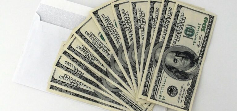 A stack of $100 U.S. dollar bills fanned out on top of a white envelope, placed on a plain white surface.