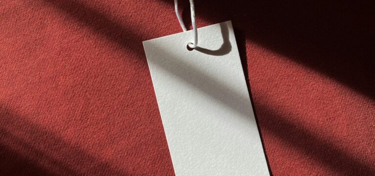 A blank price tag attached to a string rests on a burgundy fabric surface, illuminated by soft natural light and shadows.