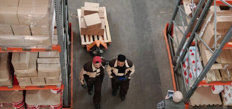 A warehouse with shelves stocked with boxes and supplies. Two workers in brown uniforms walk through the aisle, while a supervisor with a tablet observes inventory.