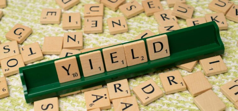 Scrabble tiles arranged to spell 'YIELD' on a green tile rack, with scattered wooden letter tiles on a textured green and white surface in the background.