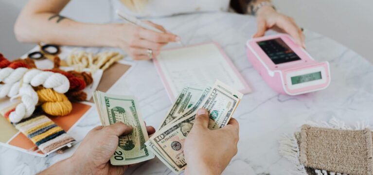 Close-up of a person counting U.S. dollar bills while another person writes in a notebook next to a pink card payment terminal. Yarn, scissors, and fabric swatches are on the marble table, suggesting a small business or craft transaction.