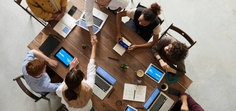 A group of diverse professionals collaborating around a large wooden table with laptops, tablets, notebooks, and smartphones. Two people in the center are shaking hands, suggesting agreement or partnership.