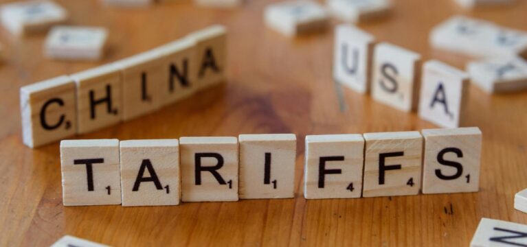 Wooden letter tiles spelling out “TARIFFS,” with blurred tiles in the background forming “CHINA” and “USA,” symbolizing international trade tensions.