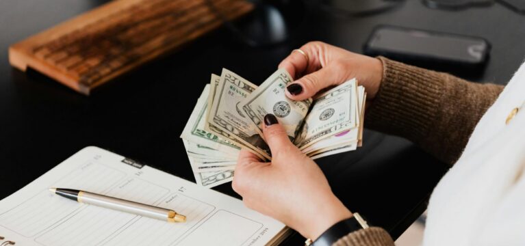A person counting U.S. dollar bills at a desk, with a planner, pen, and computer accessories nearby.