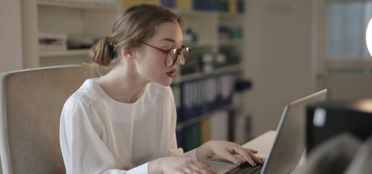 Focused businesswoman wearing glasses working on a laptop in an office, surrounded by shelves filled with organized binders and documents.