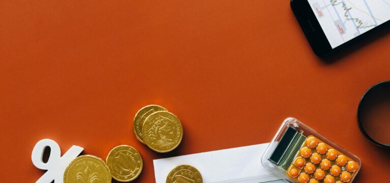 Flat lay of gold coins, a percentage symbol, a chart printout, a smartphone displaying a stock graph, and an orange calculator on a red background—symbolizing interest rates and financial analysis.