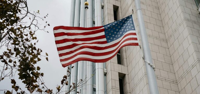 An American flag waves on a flagpole outside a modern government or corporate building with light-colored stone walls and vertical metal accents, under an overcast sky. A tree with sparse autumn leaves stands in the foreground.