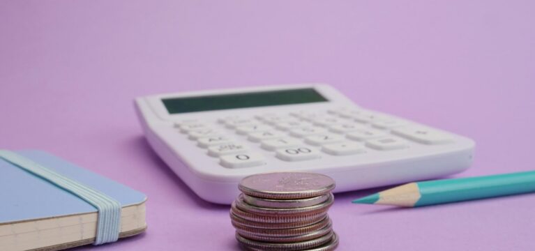 A pastel-themed flat lay featuring a white calculator, a stack of silver coins, a blue notebook with an elastic band, and a turquoise pencil with a sharpened tip, all arranged on a lavender background.