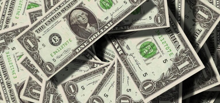 A scattered pile of U.S. one-dollar bills featuring George Washington, showing the front side of the notes with green Treasury seals and serial numbers visible.